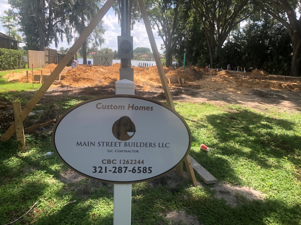 Sign for Main Street Builders LLC, a licensed contractor, in front of a construction site with earth moving and foundation markers.