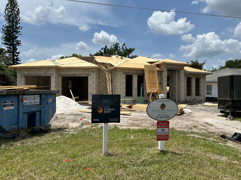Construction site of a residential building with unfinished exterior walls, wood framing, and a blue dumpster in front. Signs for Main Street Builders and a bank are visible, along with a private property warning.