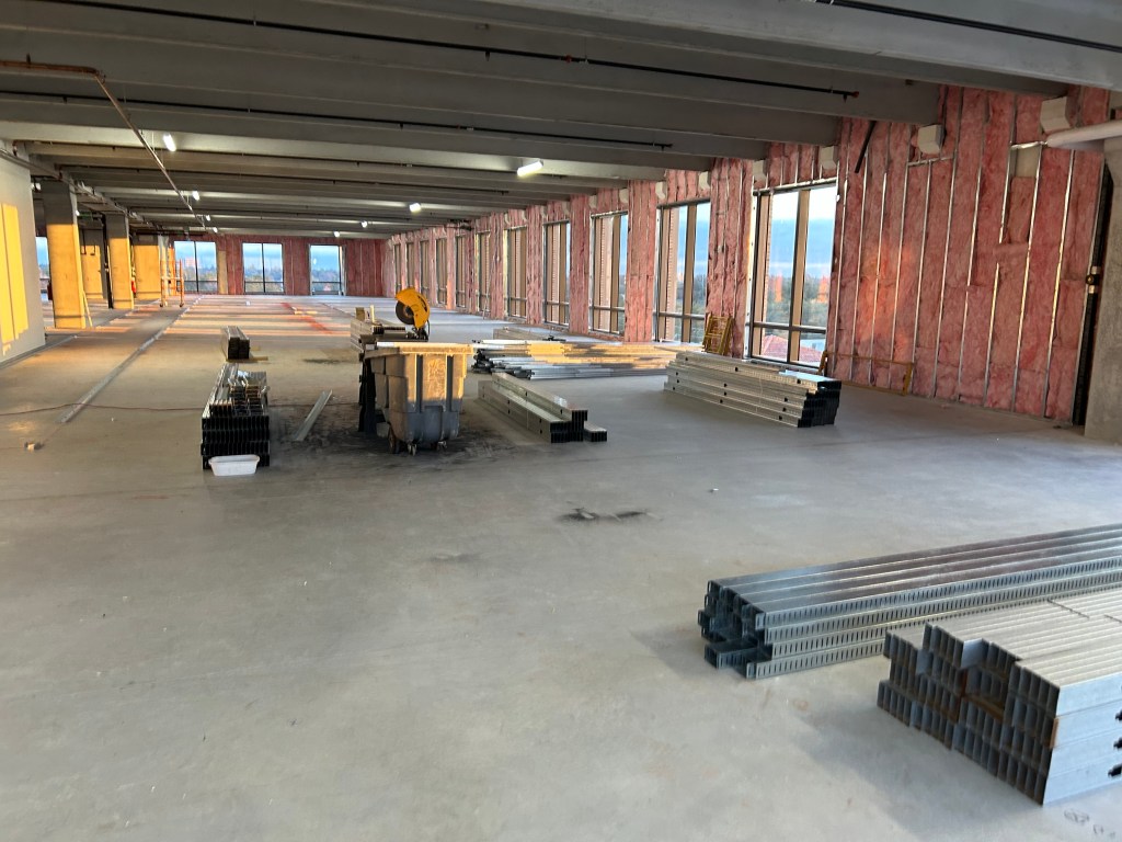 A large, unfinished commercial interior featuring concrete floors, construction materials, and exposed walls with insulation. Windows line the area, allowing natural light to enter.